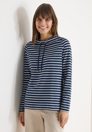 Femme souriante, portant un haut à manches longues rayé bleu marine et blanc avec des cordons, debout contre un mur blanc.