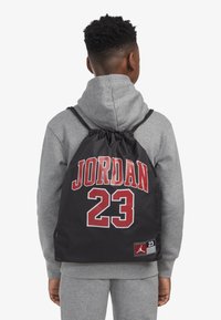 Rucsac negru cu șnur, având inscripționat în roșu textul "JORDAN" și numărul "23", realizat dintr-un material textil cu o textură netedă, purtat peste un hanorac gri.