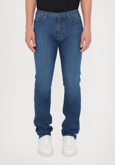 Jeans in denim blu scuro con taglio dritto. Presentano cinque tasche, chiusura frontale con bottone e cuciture a contrasto. Indossati con sneakers bianche.