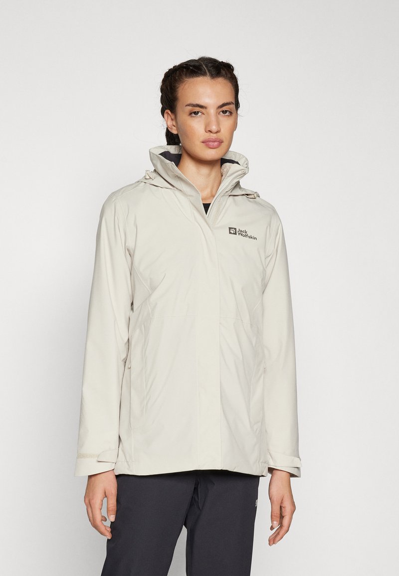 Giacca impermeabile beige chiaro con cappuccio, coulisse regolabili e un logo sul petto. Presenta una zip frontale e tasche pratiche.