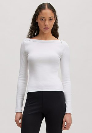 Junge Frau mit lockigem Haar, die ein eng anliegendes weißes schulterfreies Langarmshirt und schwarze Hosen trägt, stehend vor einem schlichten Hintergrund.