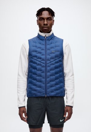 Un homme porte un gilet bleu matelassé zippé par-dessus un haut blanc à manches longues et un short de sport noir, sur un fond uni.