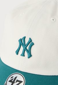 Λευκό και τιρκουάζ καπέλο μπέιζμπολ με έντονο τιρκουάζ κεντημένο λογότυπο "NY" και υφασμένο γείσο, με στρογγυλή μαύρη και λευκή ετικέτα μάρκας.