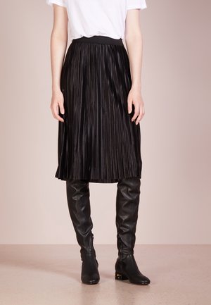 Femme portant un haut blanc à manches courtes, une jupe midi plissée noire, et des bottes en cuir noires montantes jusqu'au genou, debout devant un fond uni.