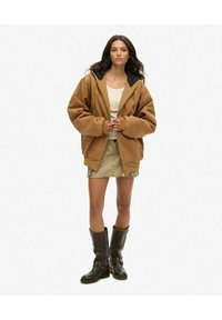 Brązowa, oversize'owa kurtka z czarnym kapturem, zestawiona z jasnobeżowym topem, khaki spódnicą i wysokimi brązowymi skórzanymi botkami z ćwiekami.