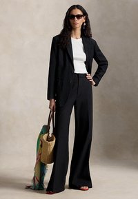 Γυναίκα που φοράει μαύρο σακάκι και παντελόνι με φαρδύ μπατζάκι, λευκό τοπ, γυαλιά ηλίου, κρατώντας ψάθινη τσάντα ώμου με πολύχρωμο μαντήλι, στέκεται μπροστά σε ουδέτερο φόντο.