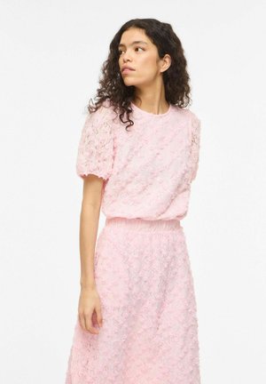 Femme aux cheveux bouclés portant un haut rose en dentelle à manches courtes et une jupe assortie, regardant vers sa gauche sur un fond blanc.