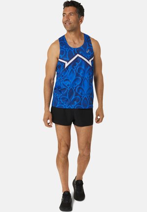 Camiseta sin mangas azul con un patrón de acabado intrincado, acentos blancos y un escote de cuello redondo. Llevada con pantalones cortos negros y zapatos deportivos.