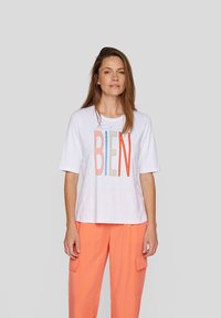 White t-shirt with multicolored text "tout est BIEN," featuring short sleeves and a relaxed fit. Paired with orange cargo pants.