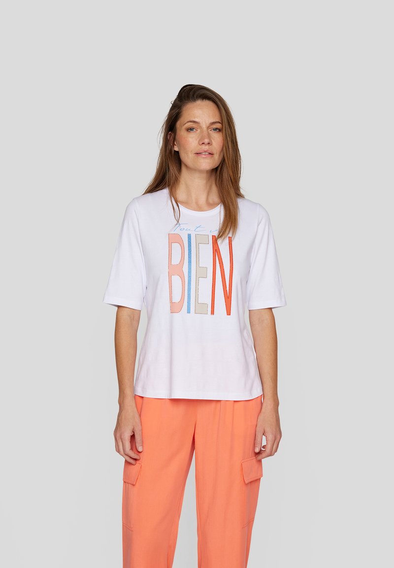 White t-shirt with multicolored text "tout est BIEN," featuring short sleeves and a relaxed fit. Paired with orange cargo pants.