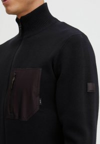 Svart zip-up genser med ribbet krage, teksturert stoff og en brystlomme med glidelås. Inkluderer et merke på ermet.