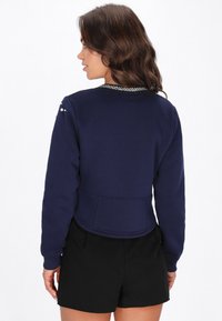 Sudadera corta azul marino con mangas largas, cuello redondo y adornos decorativos de cuentas en el hombro y el escote. Textura suave.