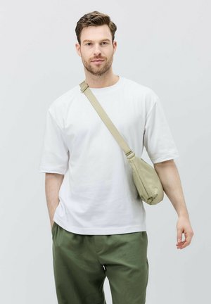 Beige skuldersæk lavet af blødt stof, båret tværs over kroppen, parret med en løs hvid t-shirt og grønne bukser. Simpelt design.