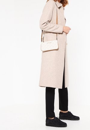 Manteau en laine beige avec un col à revers, associé à un pantalon noir et des chaussures en daim noir. Un petit sac à main blanc à motifs pend sur l'épaule.