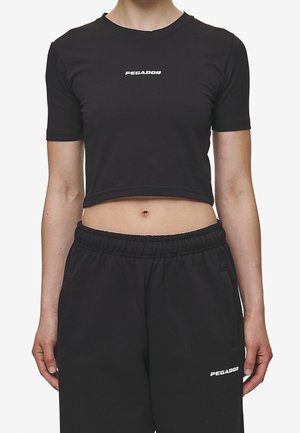 Kvinde iført sort cropped t-shirt og sorte elastiske bukser, begge med hvid "PEGADOR"-logo foran.