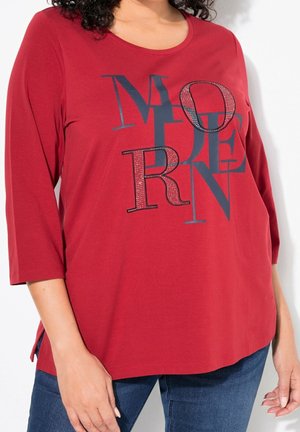 Femme portant un t-shirt rouge à manches longues avec des grandes lettres noires mélangées et cloutées sur le devant et un jean bleu, mains détendues le long du corps.