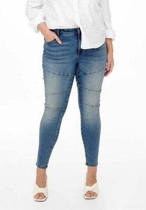 Vrouw draagt blauwe aansluitende spijkerbroek met gestikte kniepanelen, wit shirt met lange mouwen en witte open-teen hakken, staat met één hand in de zak.