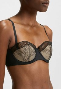 Svart bh med genomskinliga beige meshkoppar som har ett vågigt svart broderidesign, justerbara axelband och en slät underband.