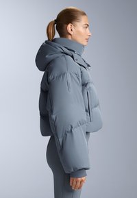 Svijetlosiva skraćena puffer jakna s kapuljačom, koja se odlikuje quilted teksturom, drukerima i detaljem bočne vrećice.