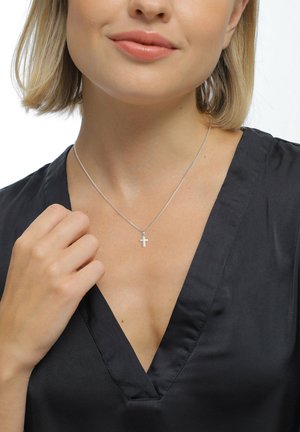 Silberne Kreuzkette an einer schlanken Kette, die auf einem schwarzen V-Ausschnitt-Oberteil ruht. Glatte, polierte Oberfläche mit einem minimalistischen Design.