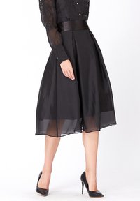 Jupe noire avec un revêtement transparent, design plissé et taille ajustée. Associée à des talons noirs, créant une silhouette sophistiquée.