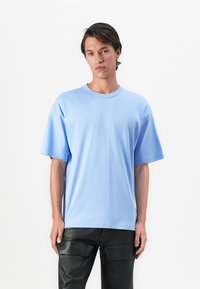 Young man wearing a light blue oversized T-shirt and black leather cargo pants, standing against a white background.