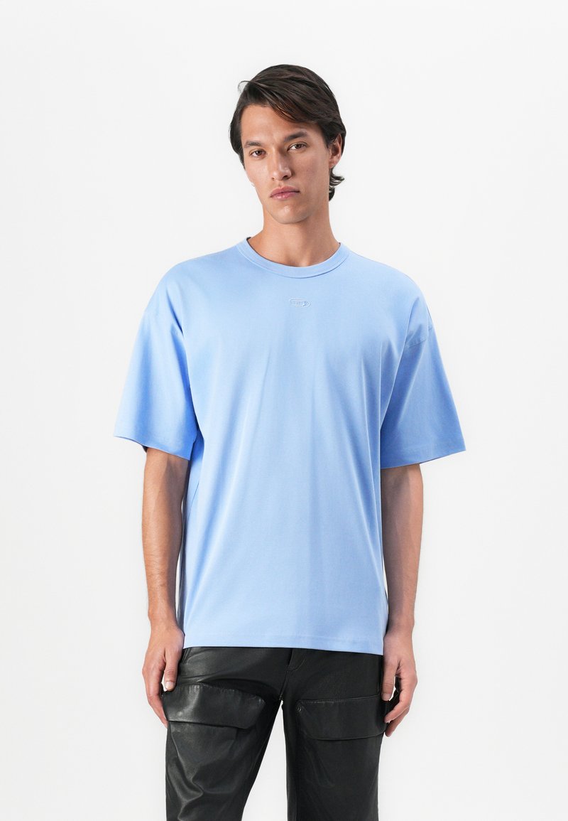 Young man wearing a light blue oversized T-shirt and black leather cargo pants, standing against a white background.