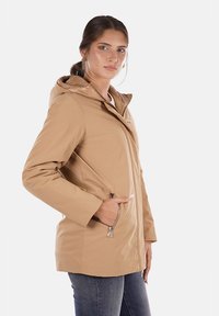 Giacca impermeabile beige con chiusura a zip, cappuccio e tasche laterali. Texture liscia e design semplice, adatta per un abbigliamento casual.