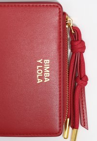 Czerwona skórzana portmonetka o teksturowej powierzchni, złote logo "BIMBA Y LOLA", suwak z plecionym sznurkiem, złote akcenty hardware.