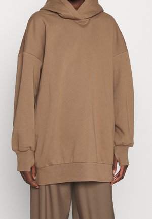 Felpa brown oversize in tessuto morbido con cappuccio, spalle scese e polsini a costine. Abbinata a pantaloni larghi coordinati.