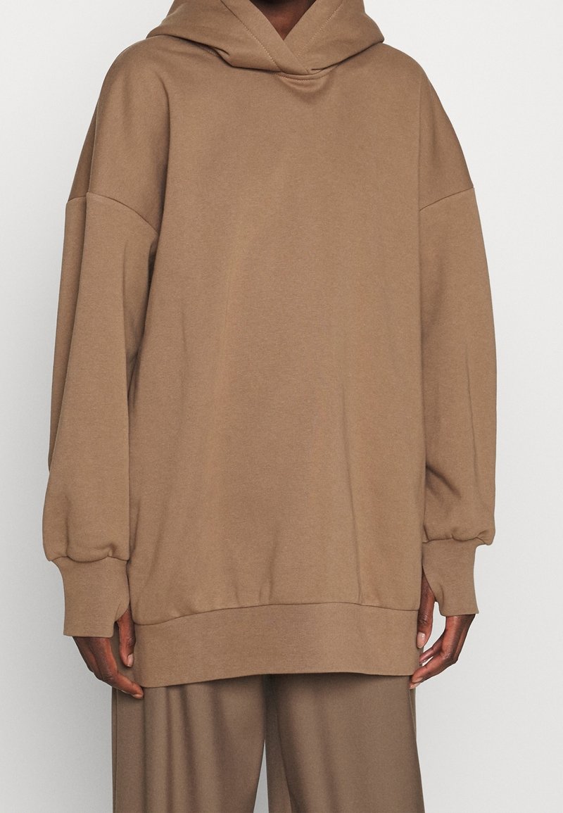 Felpa brown oversize in tessuto morbido con cappuccio, spalle scese e polsini a costine. Abbinata a pantaloni larghi coordinati.
