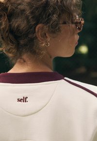 Bluzę z okrągłym dekoltem w kolorze off-white z burgundowymi akcentami. Posiada ściągacz przy dekolcie oraz haftowane logo „self.” na plecach. Gładka faktura.