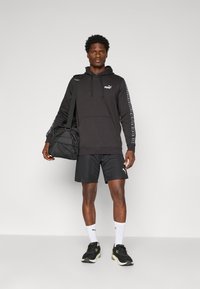 Man in zwarte Puma hoodie en shorts, witte Puma sokken, zwarte sneakers, met een zwarte sporttas over zijn schouder, staand tegen een effen achtergrond.