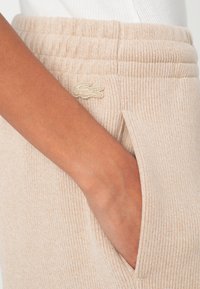 Béžové pletené tepláky s rebrovaným pásom, s bočným vreckom a malým vyšívaným logom. Mäkká textúra s jemnými pruhmi.