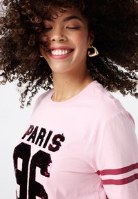 Roze katoenen t-shirt met "PARIS 96" in zwarte pailletten. Heeft twee rode strepen op de mouwen en een ronde hals. Zachte textuur.