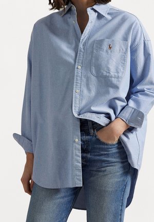 Kvinde iført en løs lyseblå skjorte med knapper, med ærmerne rullet op og en forlomme, kombineret med blå denimjeans, hånd i lommen.