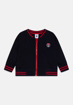 Marineblauwe ritsjack met rode strepen op de manchetten en de zoom, met een geborduurd logo op de borst en twee voorzakken. Zachte stoffen textuur.