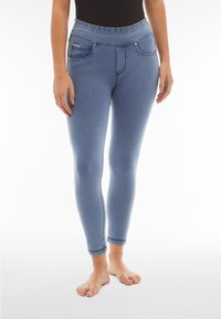 Nainen, joka käyttää vaaleansinisiä istuvia jeggingsejä etutaskuilla, seisoo paljain jaloin valkoista taustaa vasten.