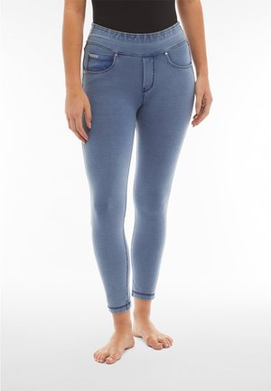 Kvinne iført lyseblå tettsittende jeggings med frontlommer, stående barfot mot en hvit bakgrunn.