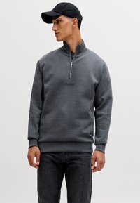 Mann trägt ein dunkelgraues Sweatshirt mit Viertelreißverschluss und eine schwarze Baseballkappe, steht mit entspannten Händen und blickt seitlich vor einem schlichten Hintergrund.