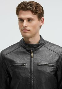 Schwarze Kunstlederjacke mit hohem Kragen, ausgestattet mit zwei Reißverschluss-Brusttaschen und einem Reißverschluss vorne, strukturiertes Oberflächendetail.