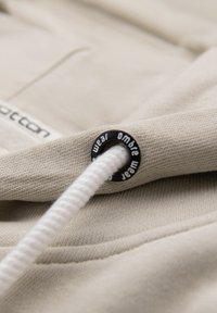 Gros plan sur un tissu beige avec un cordon blanc passant à travers un œillet en métal noir étiqueté "ombre wear".