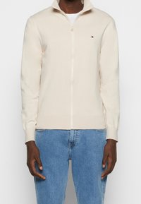 Uomo che indossa un maglione crema con cerniera e un piccolo logo Tommy Hilfiger, abbinato a jeans azzurri su uno sfondo semplice.