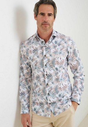 Man draagt een overhemd met lange mouwen en knopen met een pastelblauw, bruin en grijs tropisch bladpatroon, beige broek, staat met één hand in de zak.