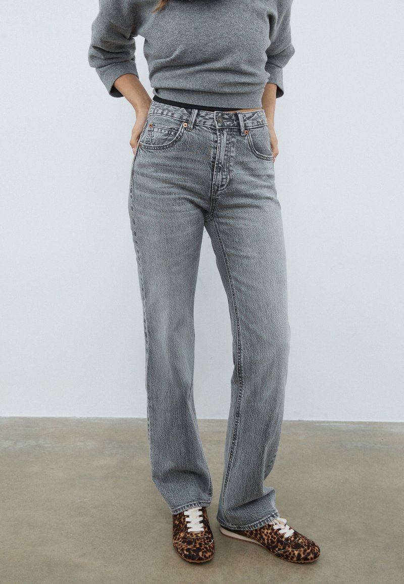 Personne portant un jean gris ample taille haute associé à un pull court gris et des baskets à motif léopard, debout sur un sol en béton.