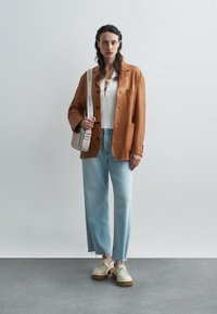 Bruine suède blazer, witte ribbel top, lichtblauwe wijde jeans en platform sneakers met beige accenten; tan crossbody tas met logoband.