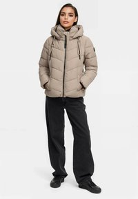 Beige gepolsterte Jacke mit strukturiertem Finish, hohem Kragen, verstellbarem Kapuzenpulli und Reißverschluss vorne, kombiniert mit schwarzen weit geschnittenen Hosen und schwarzen Schuhen.