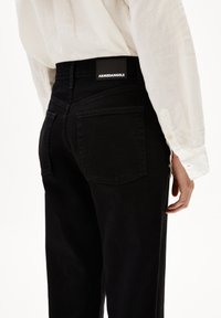 Person som bär högmidjade svarta jeans med bakfickor och en "ARMEDANGELS"-etikett, i kombination med en vit långärmad skjorta.