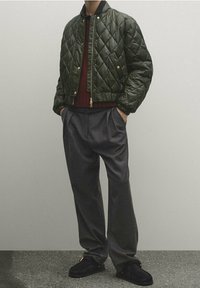 Zielona pikowana kurtka bomberka z kołnierzem, połączona z szarymi spodniami plisowanymi i czarnymi butami typu slip-on. Teksturowany i nowoczesny design.