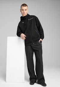 Haut à capuche noir à fermeture éclair avec un logo, comportant une capuche et des poches avant, associé à un pantalon noir ample et des chaussures noires.